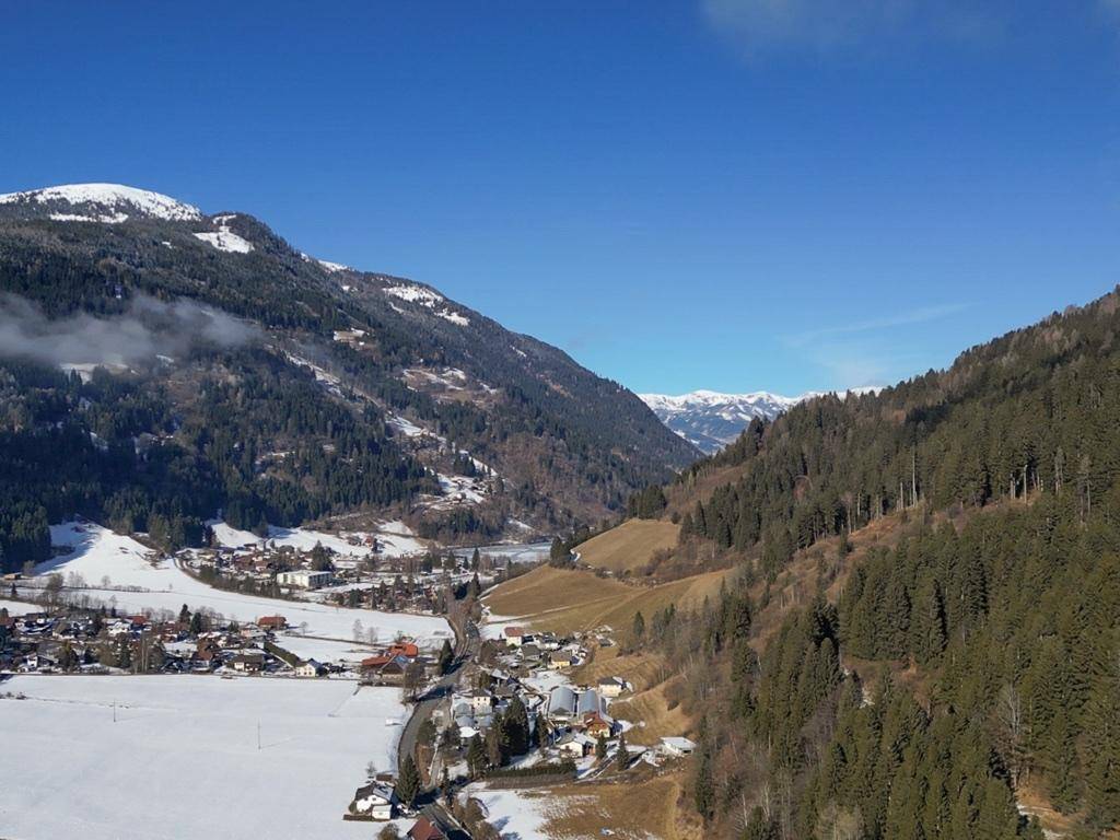 Panoramablick auf ein weites Tal mit schneebedeckten Bergen, Wäldern und einer kleinen Siedlung.