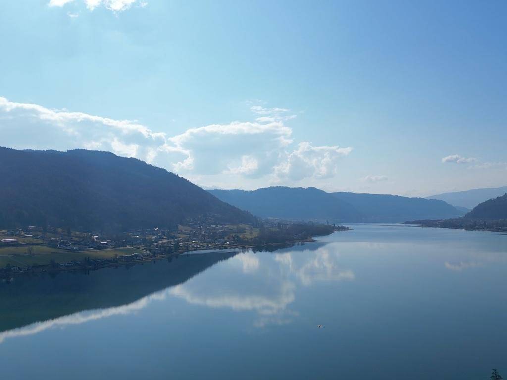 Weitläufiger Blick über einen ruhigen See, umgeben von bewaldeten Bergen unter blauem Himmel.