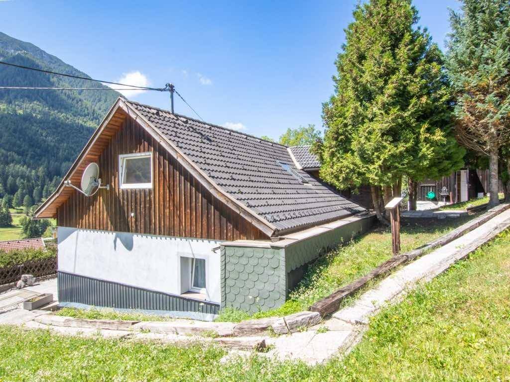 Haus mit Satteldach, Holzverkleidung und Satellitenschüssel, umgeben von grüner Landschaft.