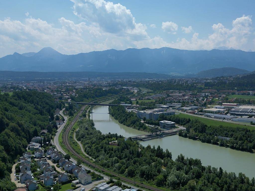 Panoramablick auf eine Stadt mit Fluss, Brücke und Bergen im Hintergrund unter blauem Himmel.