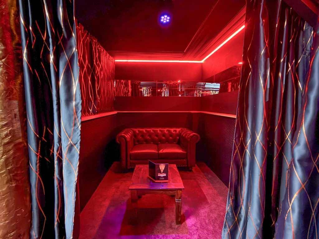 Privater Lounge-Bereich mit einem Ledersofa, Couchtisch und roten Neonlichtern, abgetrennt durch Vorhänge.