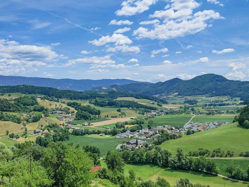 Weitläufiger Panoramablick über grüne Täler, Felder und kleine Siedlungen unter blauem Himmel.