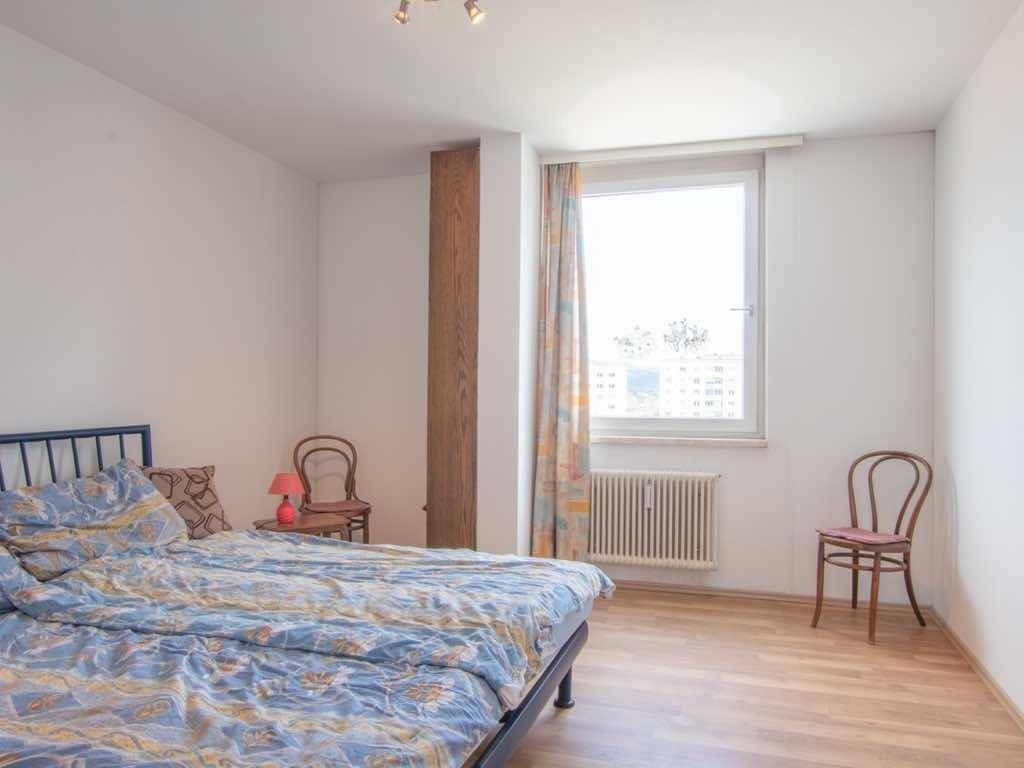Schlichtes Schlafzimmer mit Bett, Holzboden und einem Fenster, das viel Tageslicht hereinlässt.