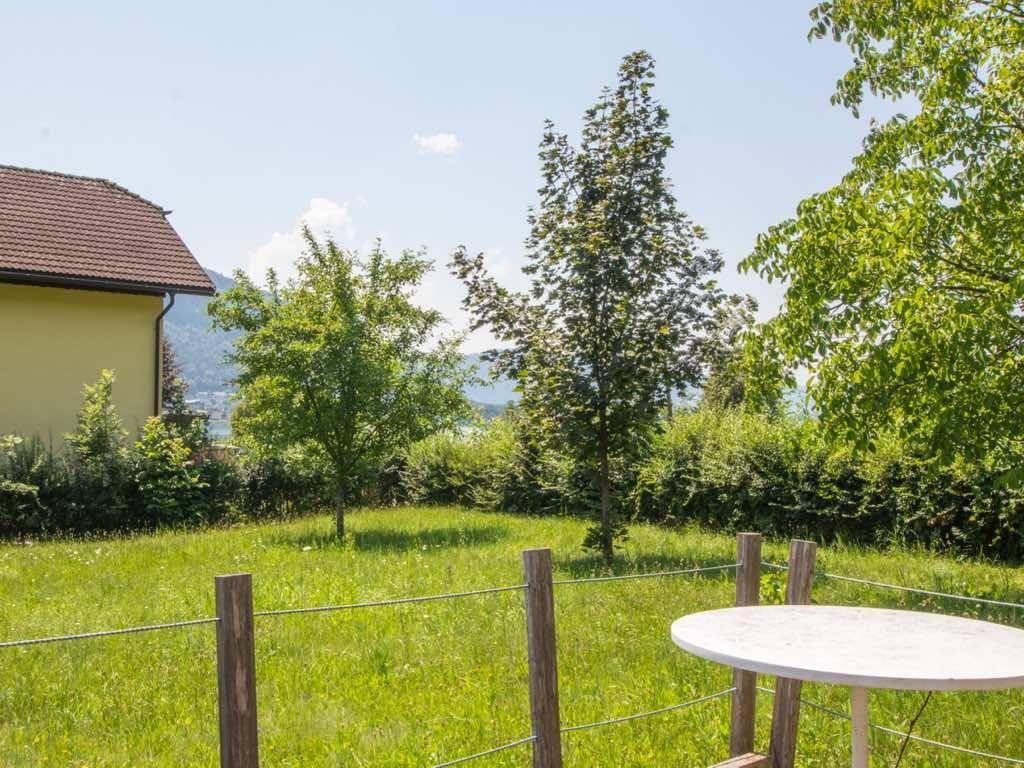Gepflegte Rasenfläche mit Gartentisch und Zaun, im Hintergrund ein Haus und Seeblick.