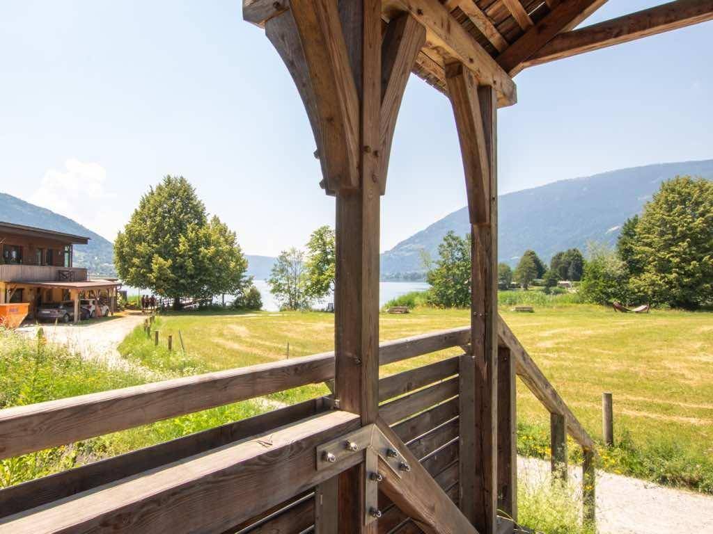 Blick von einer überdachten Holzkonstruktion auf eine grüne Wiese, den See und die Berge.