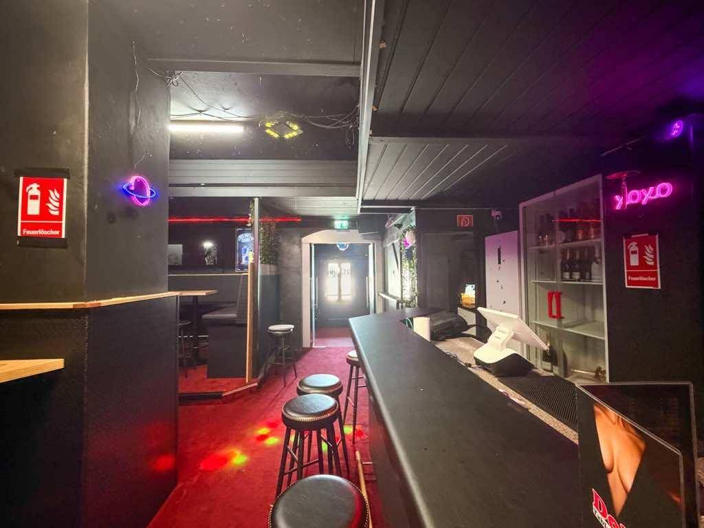 Barbereich mit Theke, Hockern und rotem Teppichboden, beleuchtet von Neonlichtern und Discokugeln.