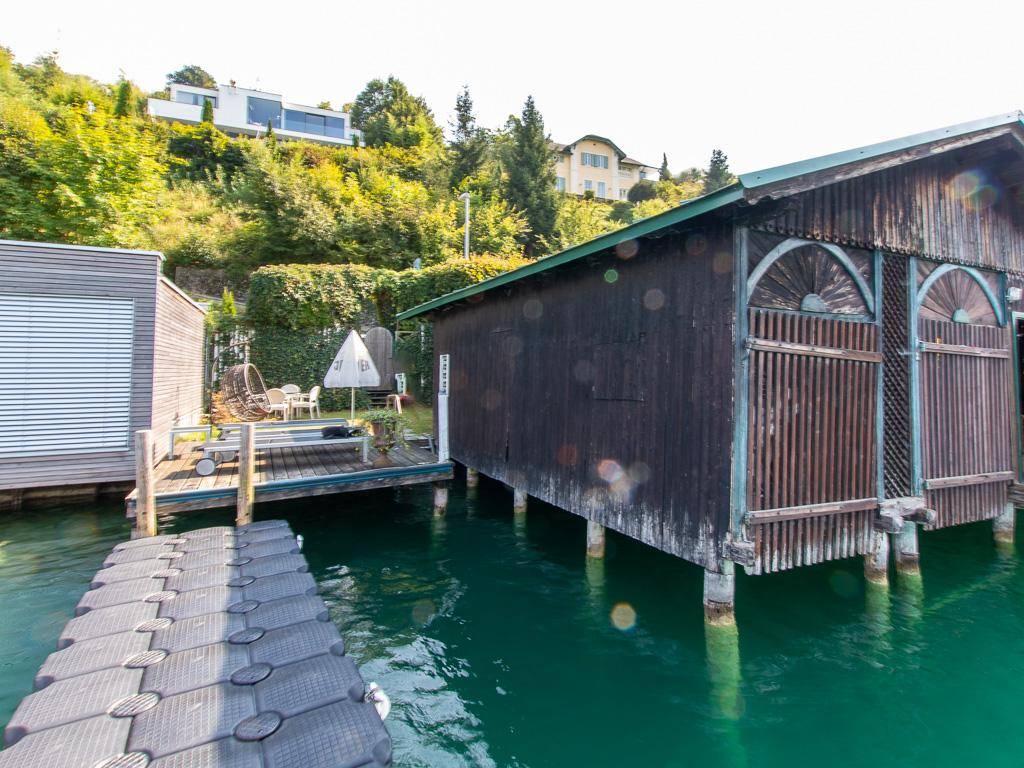 Traditionelles Holzbootshaus am See mit schwimmendem Steg und grünen Hügeln im Hintergrund.