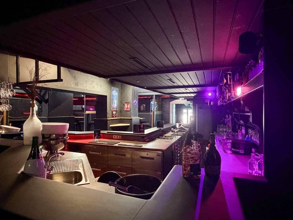 Bartresen mit Flaschen und Gläsern, beleuchtet von violettem Licht, im Hintergrund weitere Sitzbereiche.