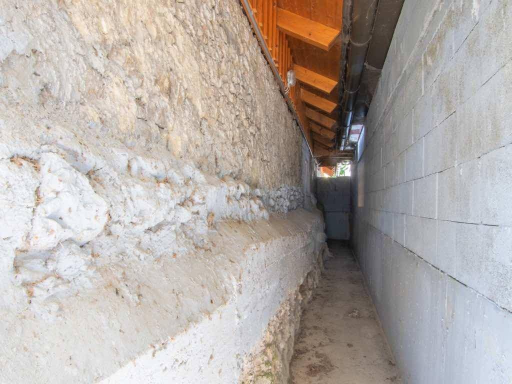 Schmaler Durchgang zwischen einer Steinmauer und einer Betonwand, der zu einer Tür führt.
