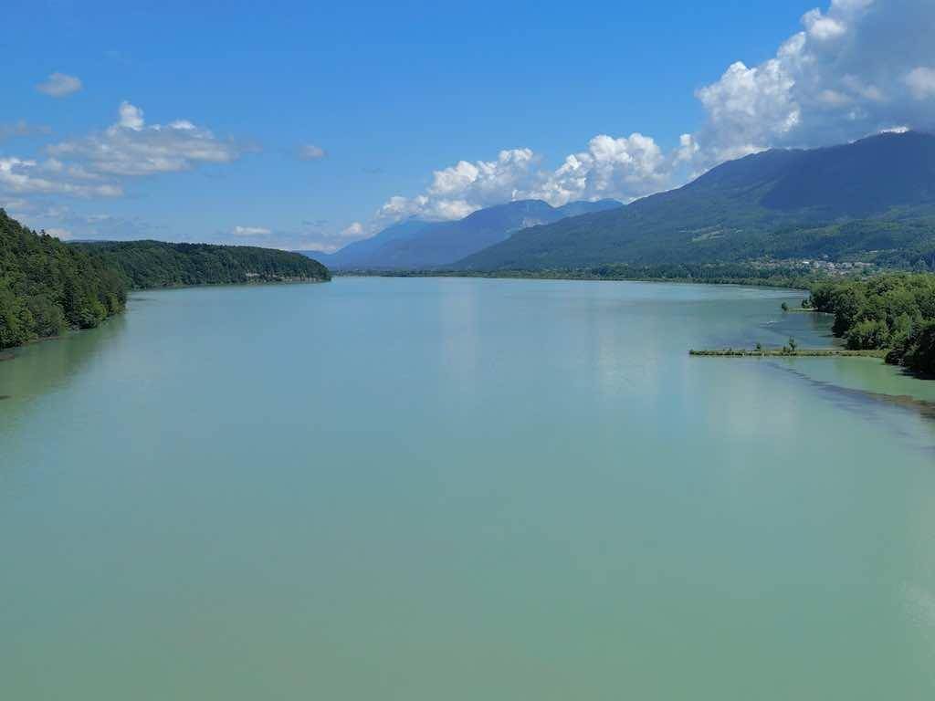 Weitläufiger Blick auf einen ruhigen See, eingebettet in eine Landschaft aus Bergen und Wäldern unter blauem Himmel.