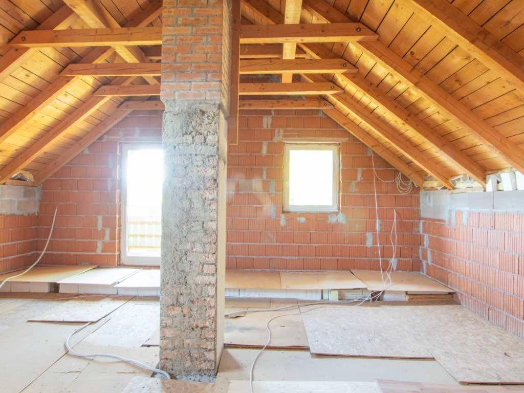 Unausgebauter Dachboden mit sichtbaren Holzbalken, Ziegelwänden und Fenstern, bereit zum Ausbau.