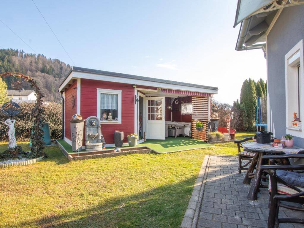 Rotes Gartenhaus mit kleiner Terrasse und Sitzgelegenheiten im sonnigen Garten.