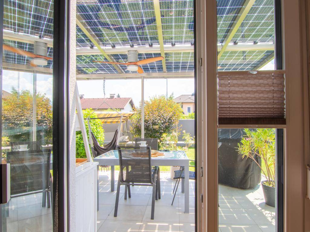 Blick vom Innenraum auf die überdachte Terrasse mit Solarpaneelen und Garten.
