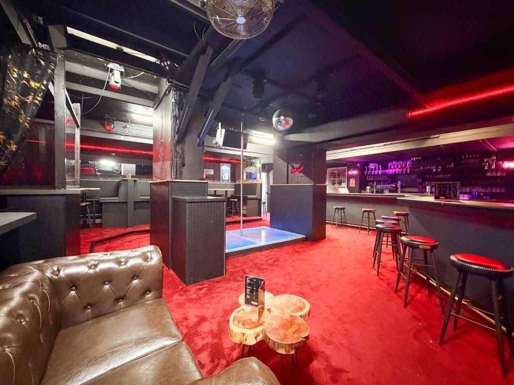 Großer Barbereich mit Tanzstange, Ledersofa, Hockern und rotem Teppichboden unter Discokugeln.