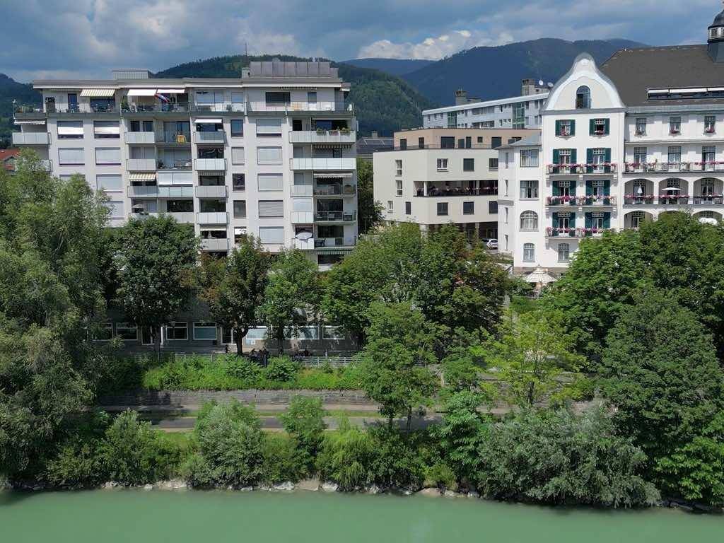Blick auf Gebäude entlang eines Flusses, umgeben von grünen Bäumen und Bergen im Hintergrund.