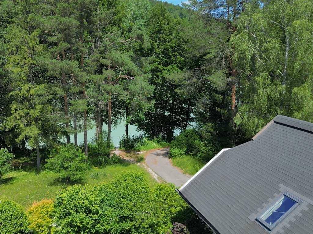 Blick über ein Hausdach mit Dachfenster auf einen See, umgeben von dichten Wäldern und grüner Landschaft.