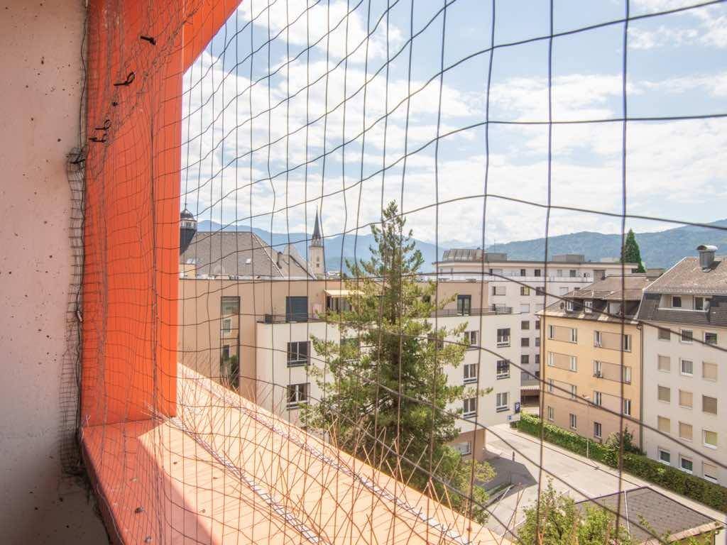 Weitblick über die Stadt mit Kirchturm und Bergen im Hintergrund, durch ein Schutznetz gesehen.