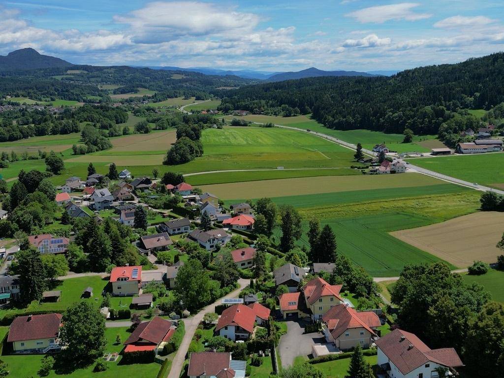 Weitläufige Luftaufnahme eines Dorfes in einer grünen Landschaft mit Feldern, Wäldern und Bergen.