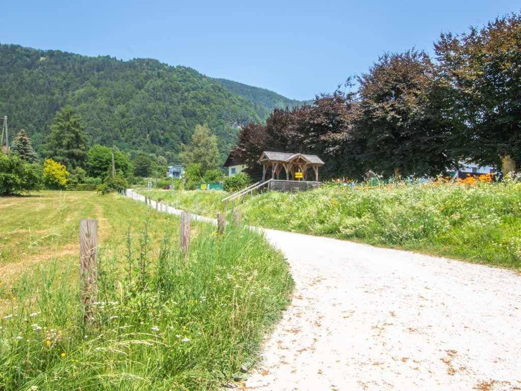 Idyllischer Schotterweg durch eine blühende Wiese, der zu einer kleinen Holzbrücke führt.