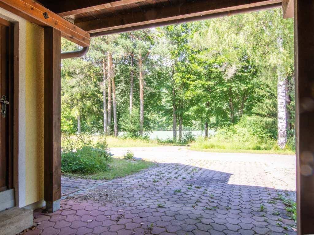 Überdachter Eingangsbereich mit gepflastertem Vorplatz und Blick auf die umliegende Natur und einen See.