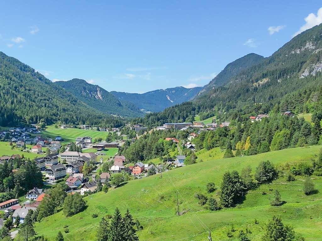 Panoramablick auf ein malerisches Dorf in einem grünen Tal, umgeben von bewaldeten Bergen unter blauem Himmel.