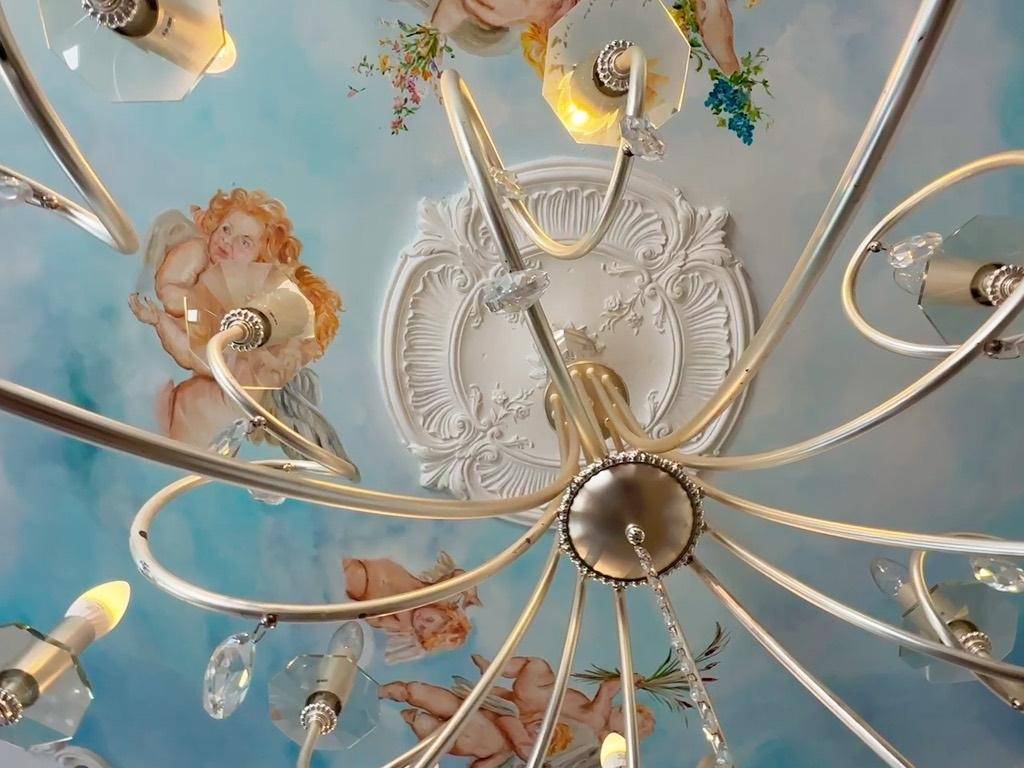 Detailansicht eines Kronleuchters vor einer kunstvoll bemalten Decke mit Stuckelementen.