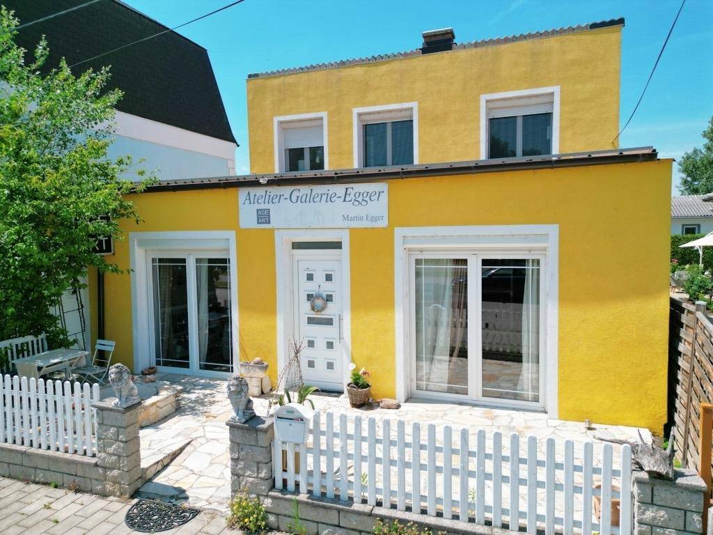 Gelbe Hausfassade mit weißen Fenstern und Türen, umgeben von einem kleinen Vorgarten mit Zaun.