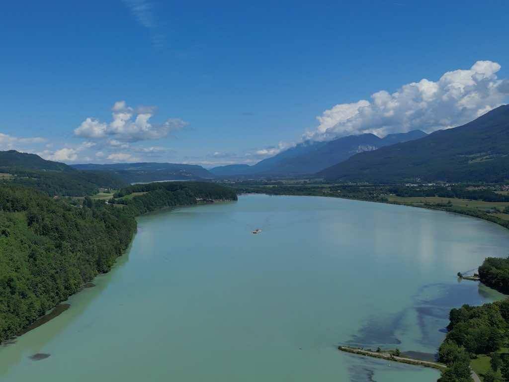Panoramablick auf einen ruhigen See, umgeben von grünen Wäldern und majestätischen Bergen unter blauem Himmel.