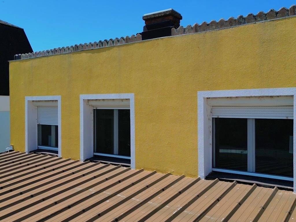 Gelbe Hausfassade mit drei Fenstern und einem braunen Wellblechdach.