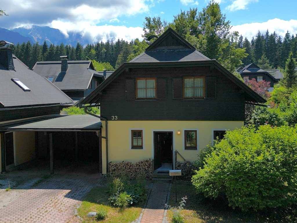 Charmantes Haus mit Holzfassade und Carport, umgeben von Bäumen und mit Bergblick.