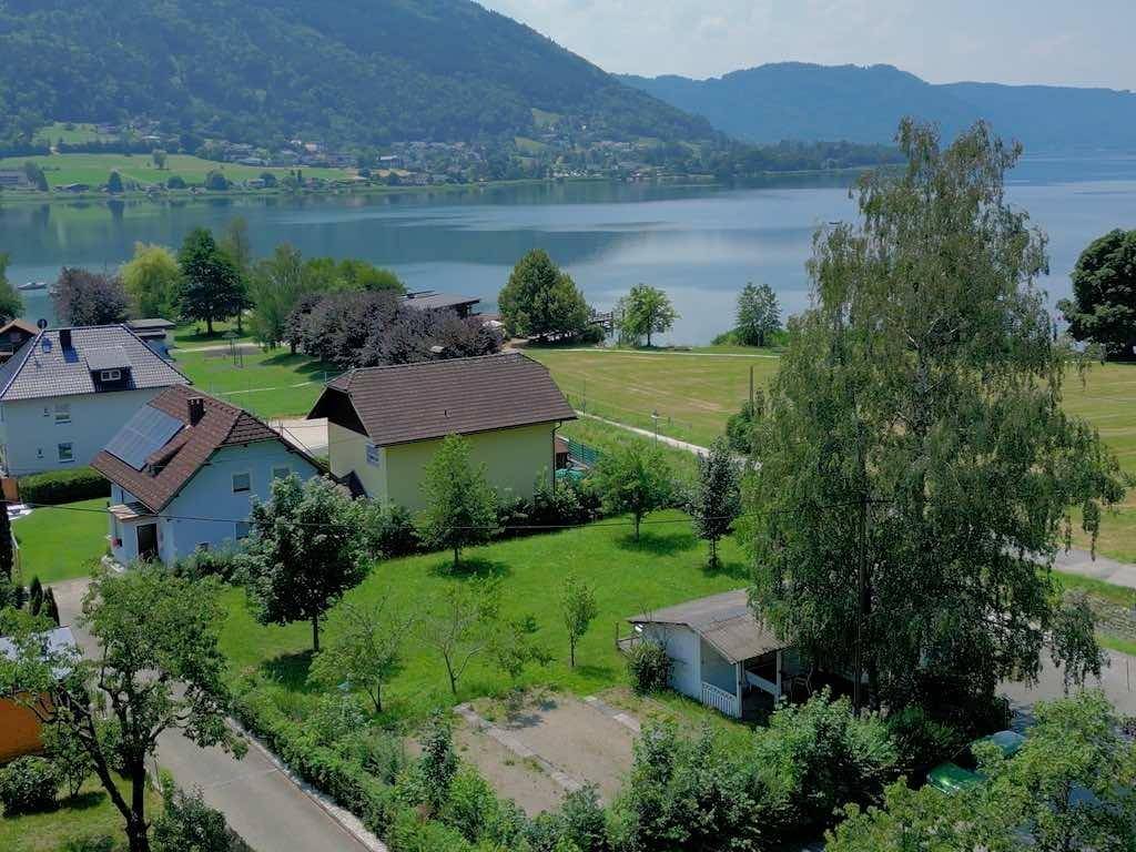 Luftaufnahme von Wohnhäusern am Seeufer mit üppiger Vegetation und Bergpanorama.