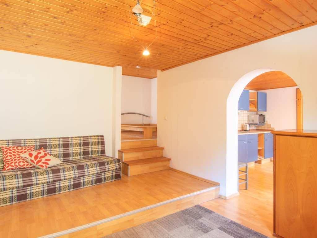Wohnzimmer mit kariertem Sofa, Holzdecke und einem Durchgang zur Küche, mit Treppe im Hintergrund.