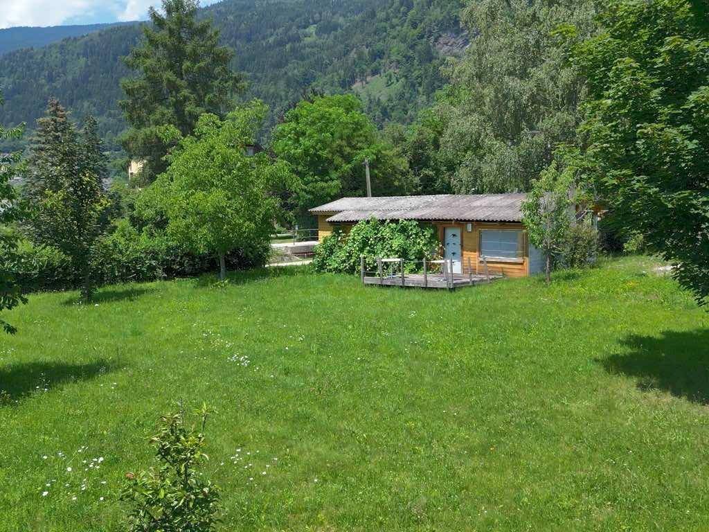 Kleines Holzhaus inmitten einer weitläufigen Wiese, umgeben von Bäumen und Bergen.