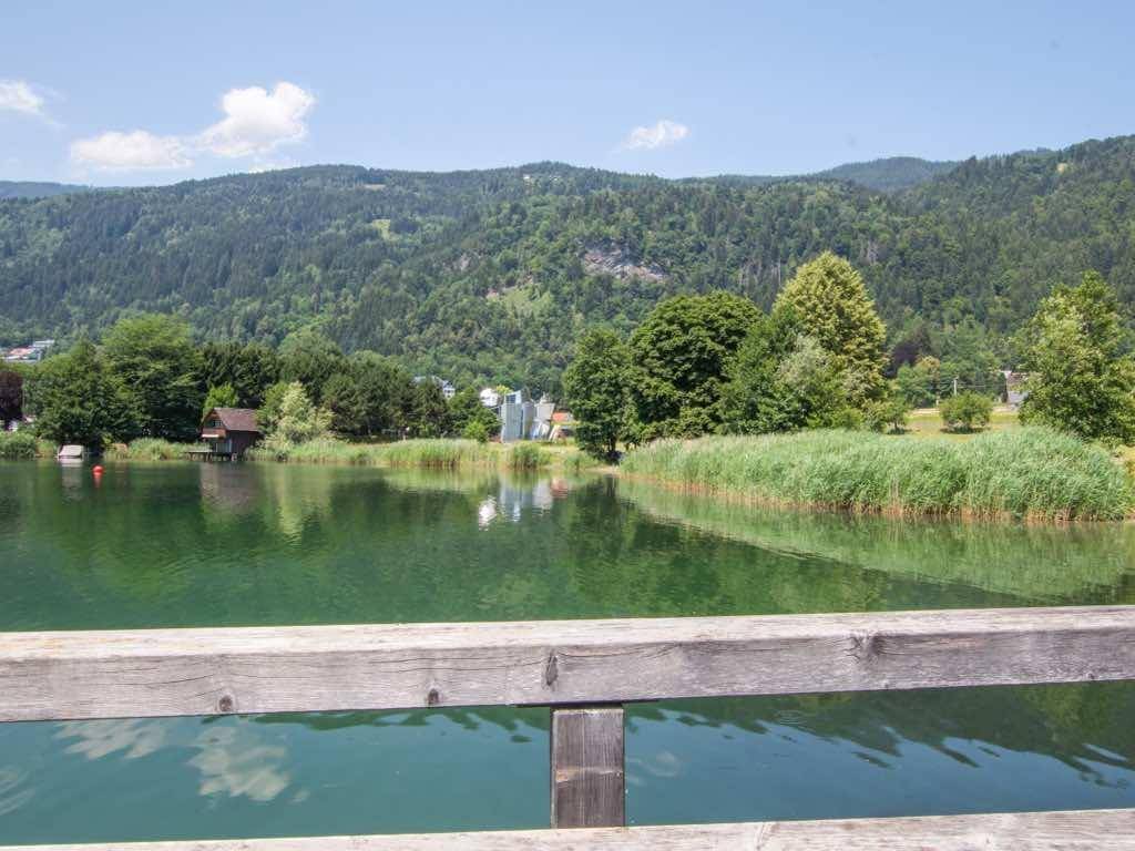 Blick über den See mit Schilfgürtel, im Hintergrund Häuser und bewaldete Berge.