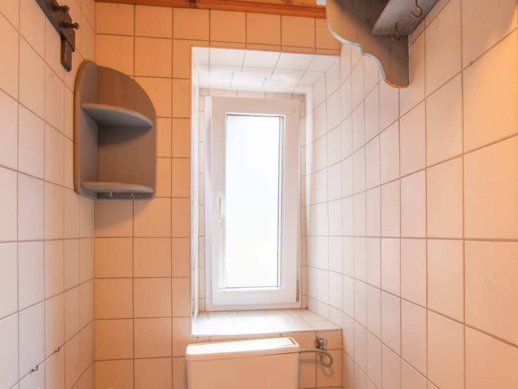 WC-Raum mit weißen Fliesen, Toilette und einem schmalen Fenster unter einer Holzdecke.