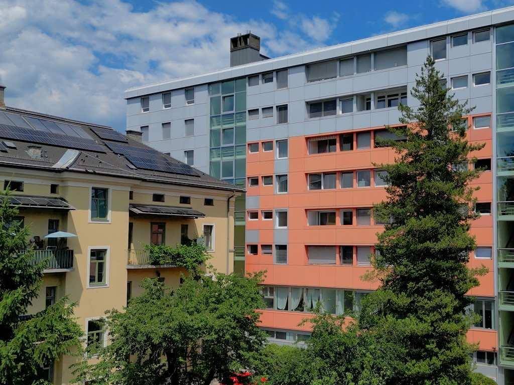 Ansicht des modernen Gebäudes mit farbigen Fassadenplatten und hohen Bäumen im Vordergrund.