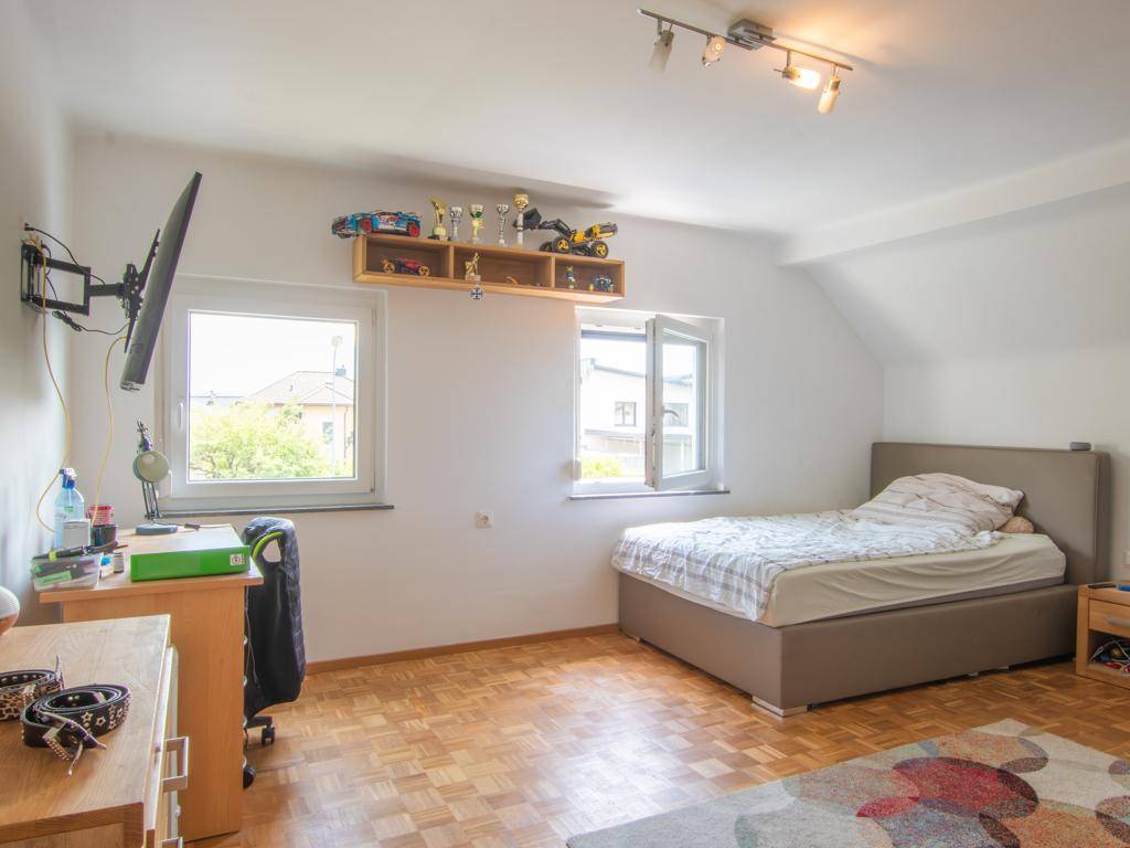 Helles Schlafzimmer mit Bett, Schreibtisch und Fenstern, möglicherweise im Dachgeschoss.