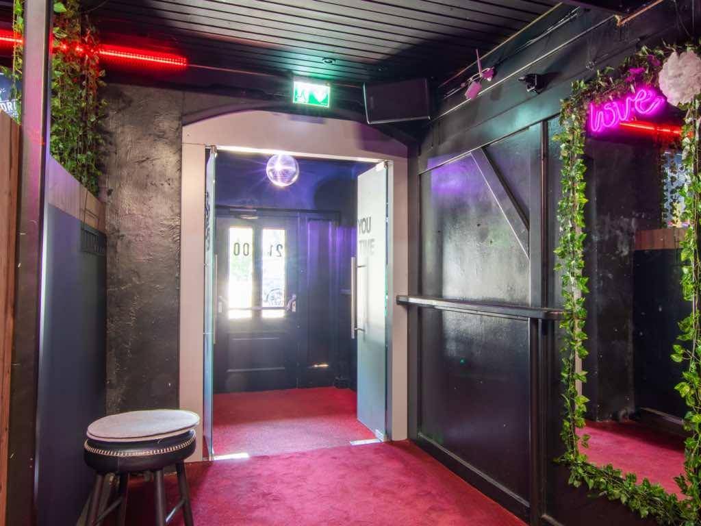 Eingangsbereich einer Bar mit rotem Teppich, grünen Ranken und einem Discokugel-Lichteffekt.