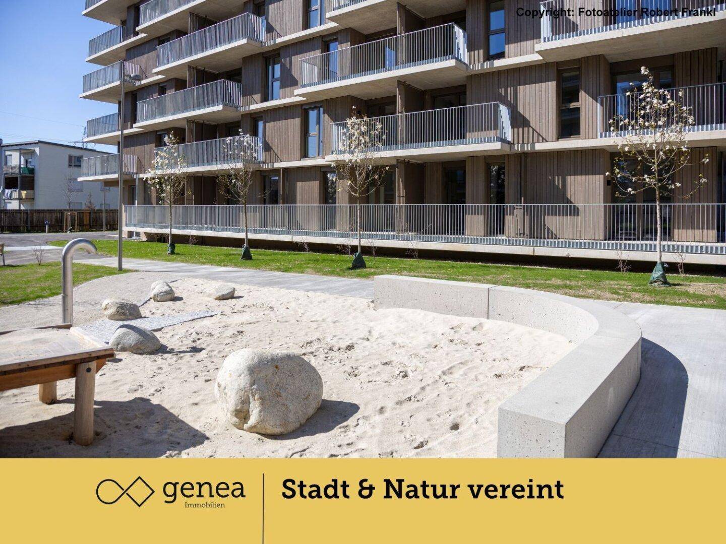 Moderner Spielplatz mit Sandbereich und Felsen vor einem neuen Wohngebäude mit Holzfassade.