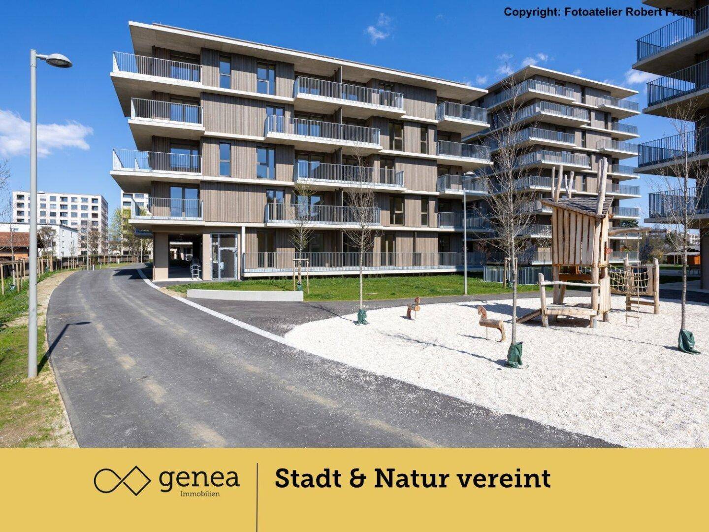 Spielplatz mit Holzklettergerüst und modernen Wohngebäuden im Hintergrund an einem sonnigen Tag.