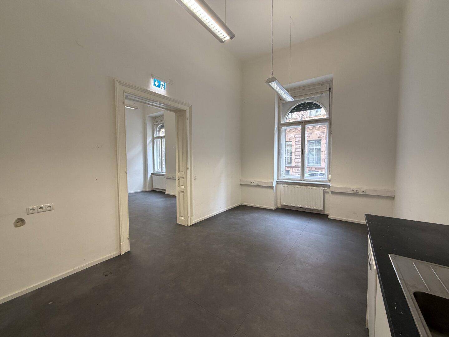 Büroraum mit Küchenzeile und Zugang zu weiteren Räumen, hell durch Fenster.