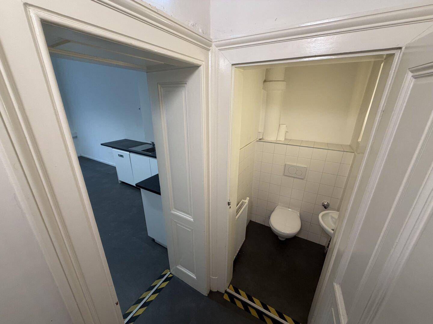 Eingangsbereich zu einem WC mit Toilette und Waschbecken, angrenzend an einen Büroraum.