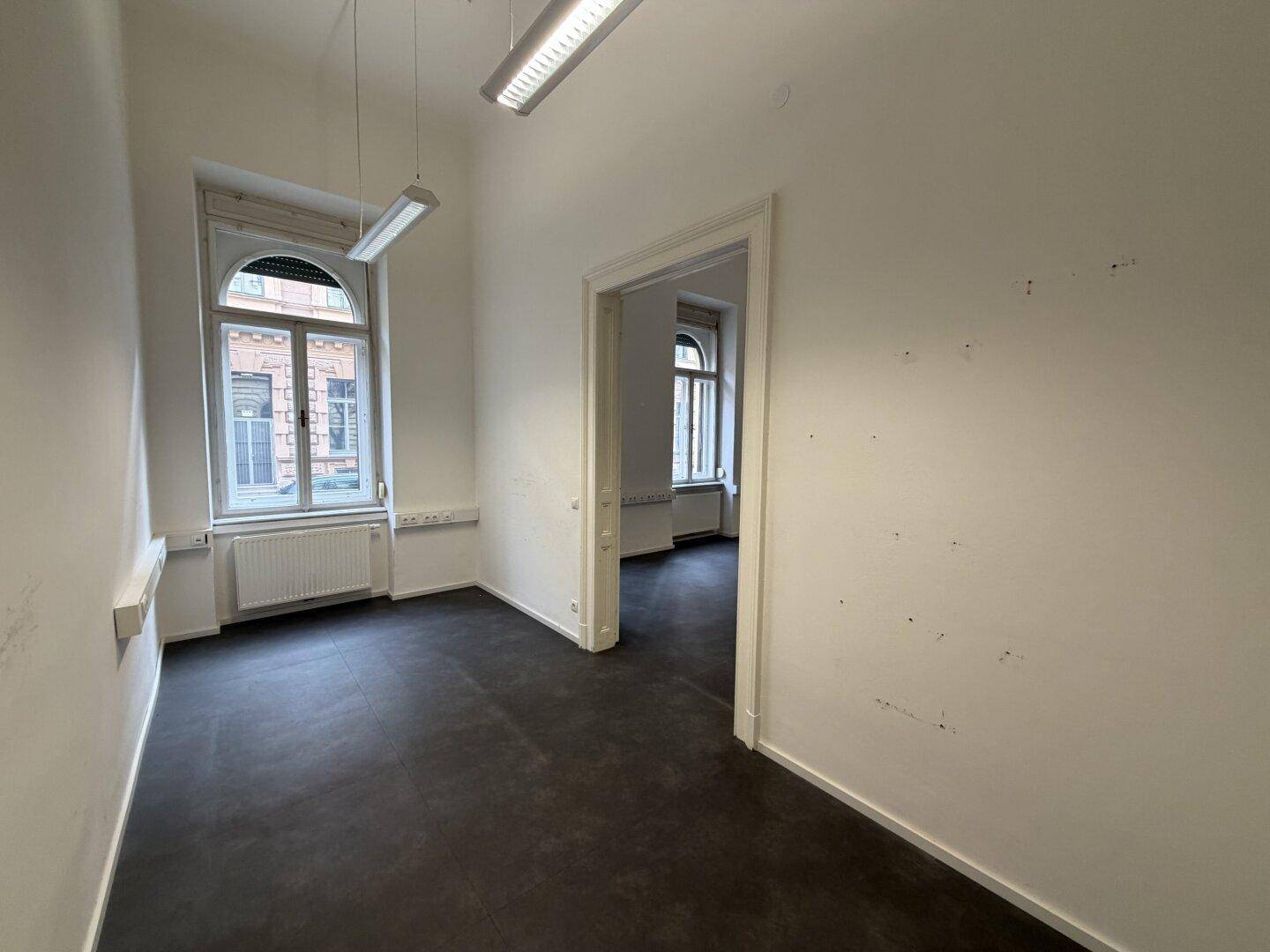 Helles Büro mit hohen Fenstern und Zugang zu weiteren Räumen, ideal für Büronutzung.