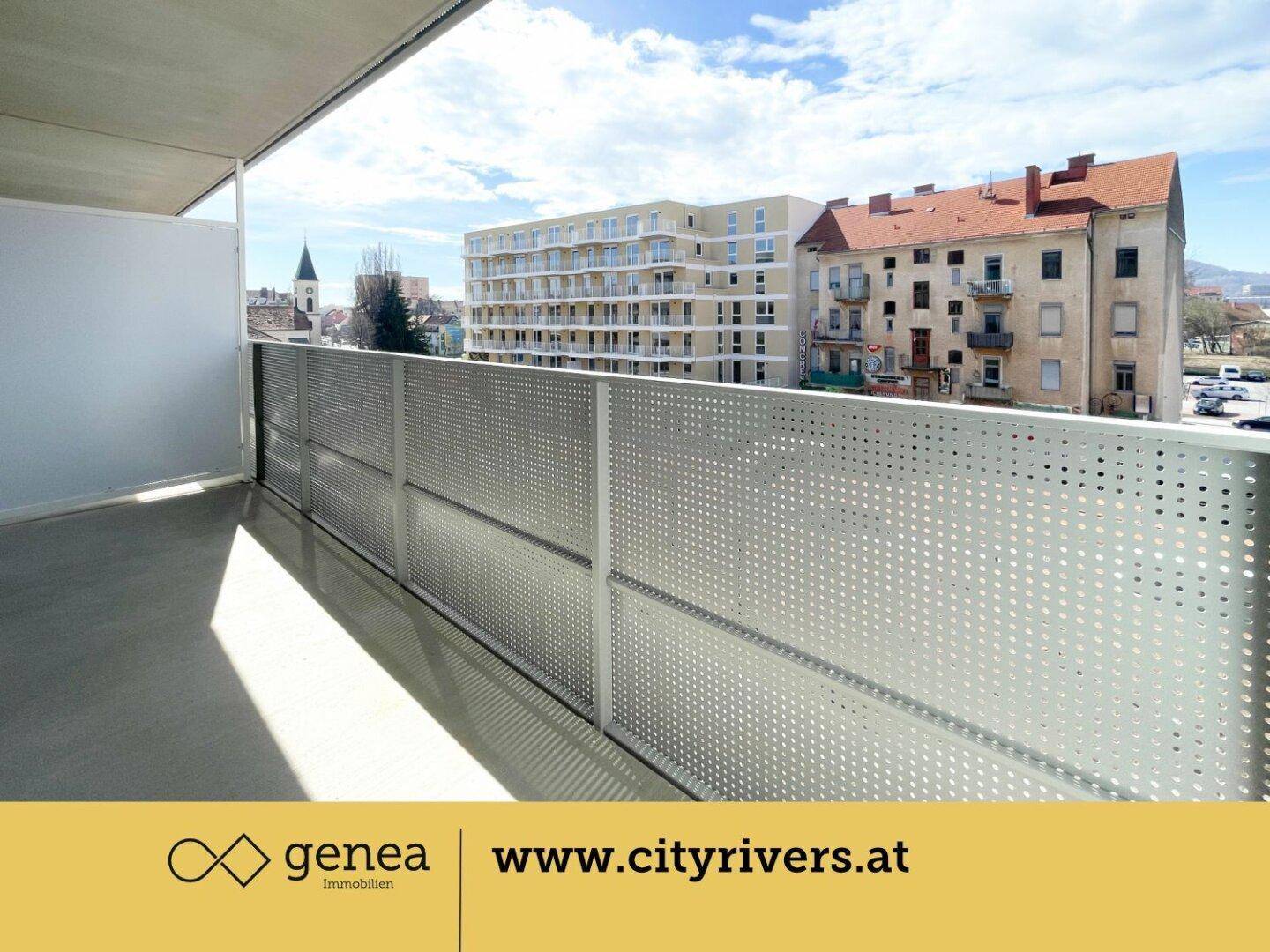Heller Balkon mit perforierter Metallbrüstung und Ausblick auf die Stadt und einen Kirchturm.