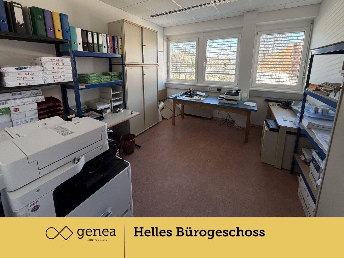 Büro mit Regalen voller Akten und einem Drucker, große Fenster sorgen für ausreichend Tageslicht am Arbeitsplatz.