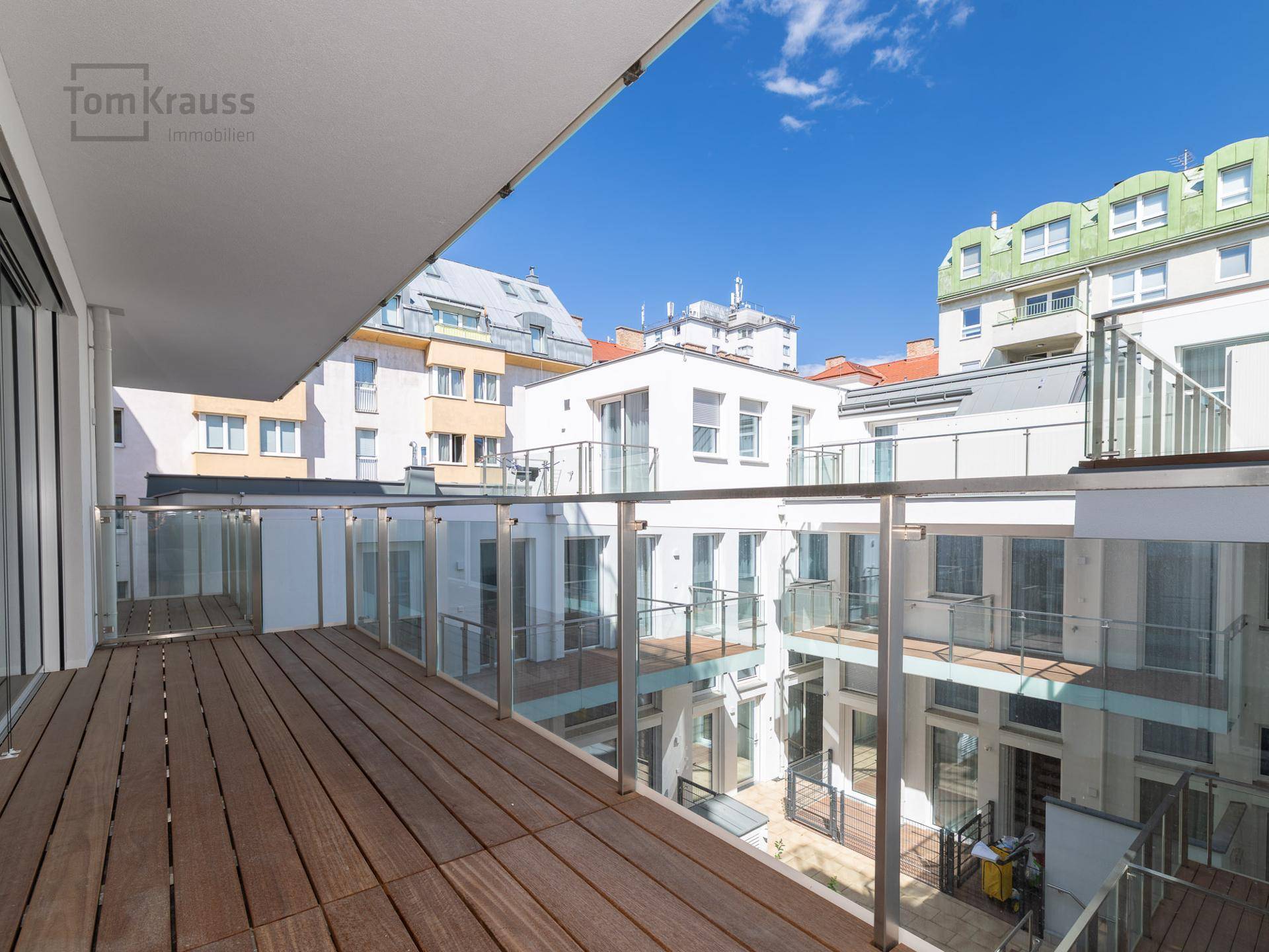 Geräumiger Balkon mit Holzbelag und Glasgeländer, bietet einen weiten Blick auf die umliegenden Gebäude.