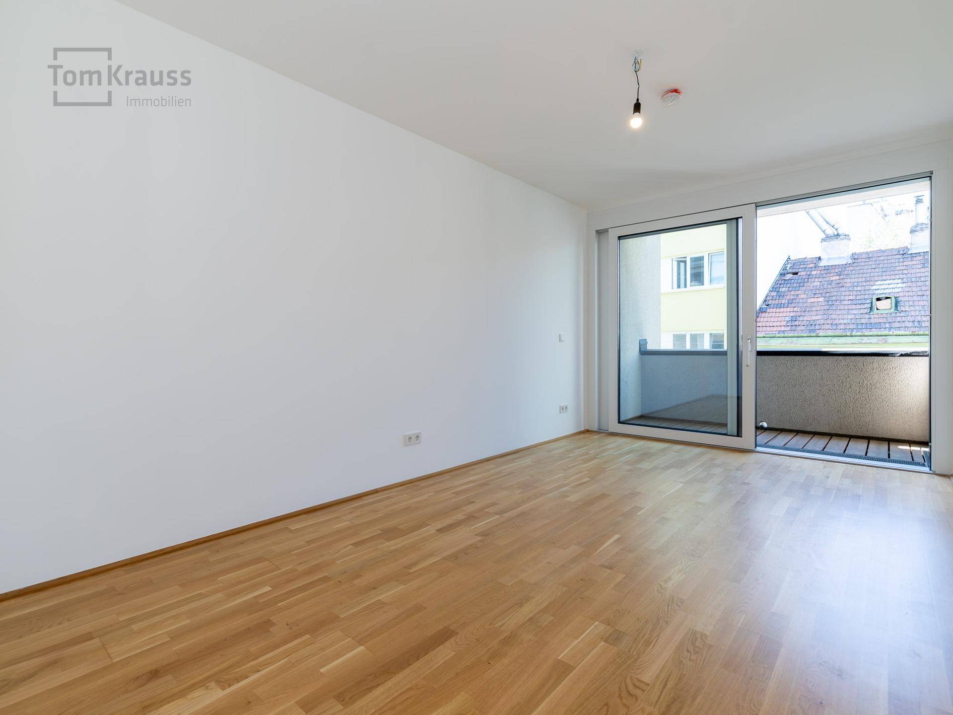 Leerer Raum mit hellem Parkettboden und großen Schiebetüren, die zu einem Balkon führen.
