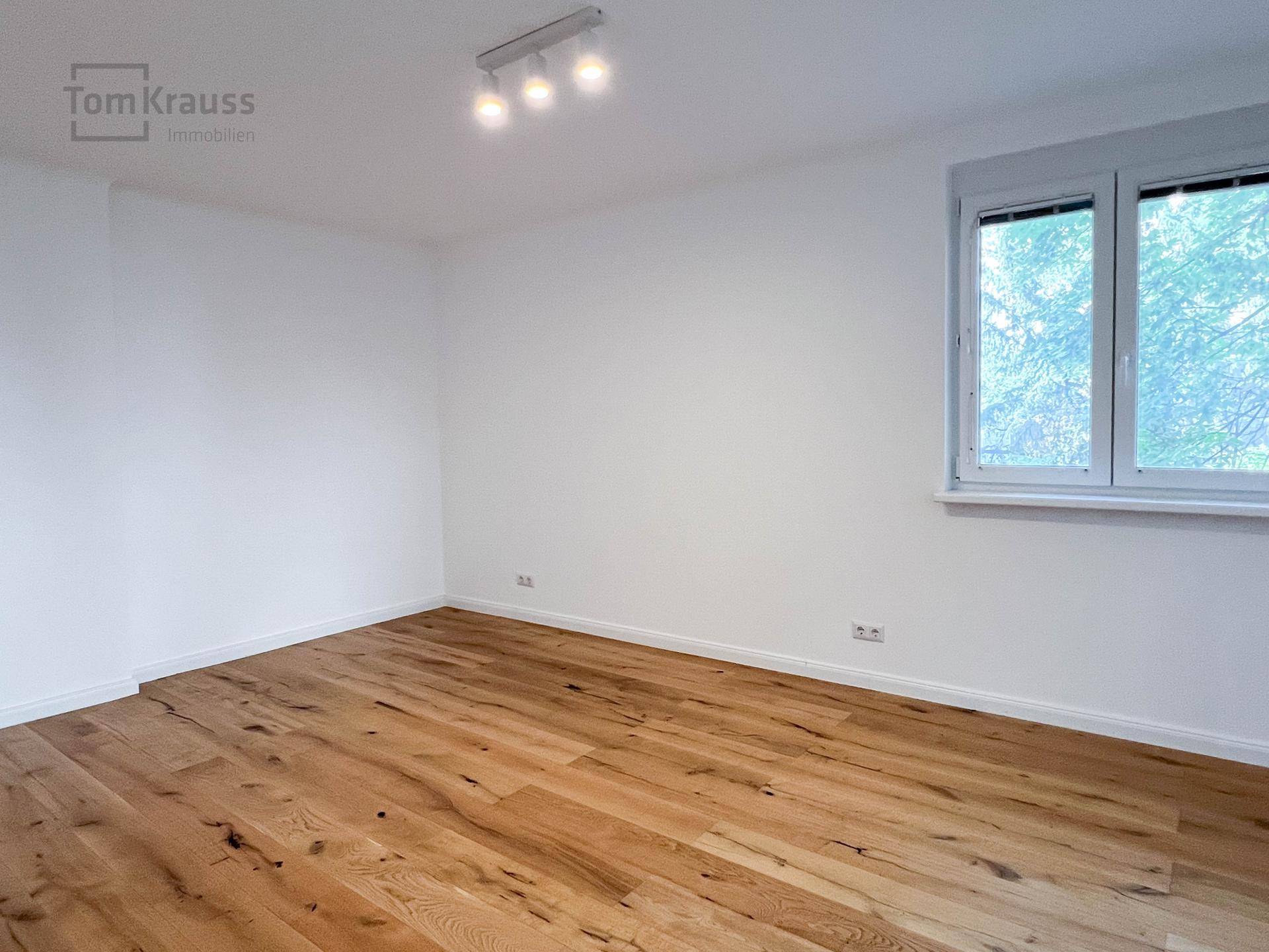 Geräumiges Zimmer mit Holzboden, weißen Wänden und großem Fenster.