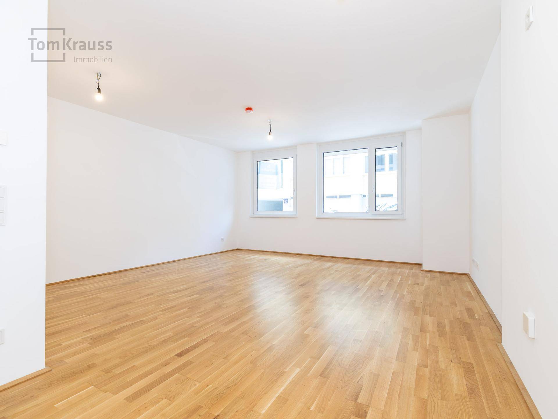Geräumiges, leeres Wohnzimmer mit hellem Parkettboden und zwei großen Fenstern.