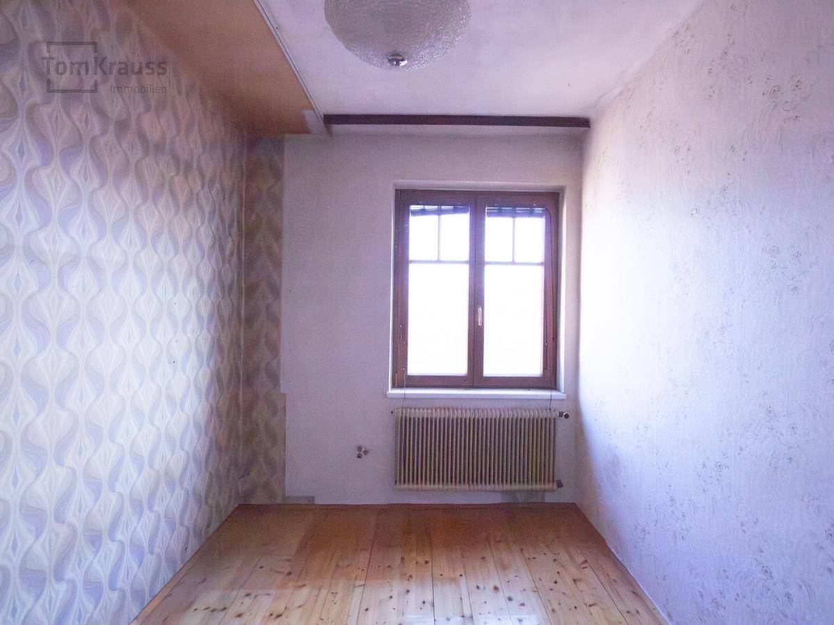 Leeres Schlafzimmer mit Holzdielenboden, gemusterter Tapete und einem Fenster mit Heizkörper.